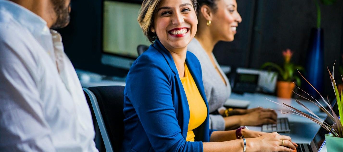Three diverse professionals working and smiling at office desks, fostering teamwork and collaboration.